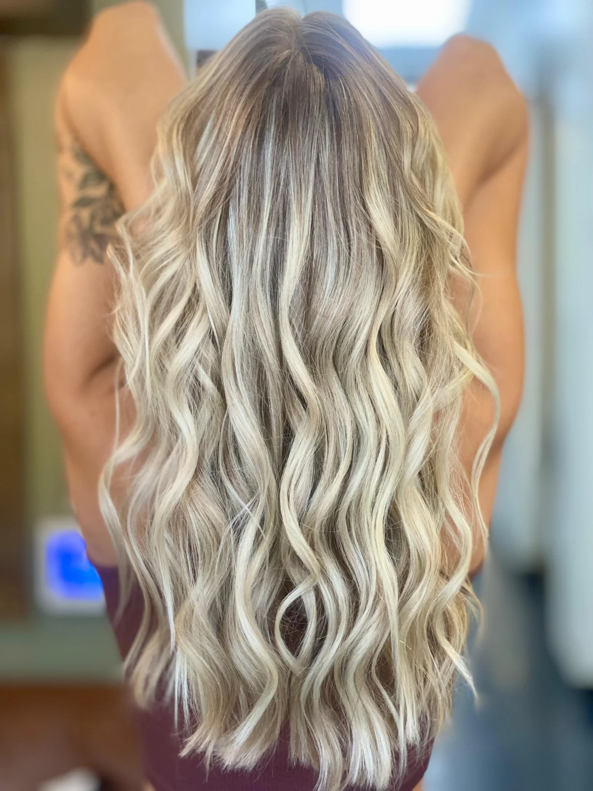 Long beige-blonde waves with brown root depth, shown from the back against a softly blurred salon background.
